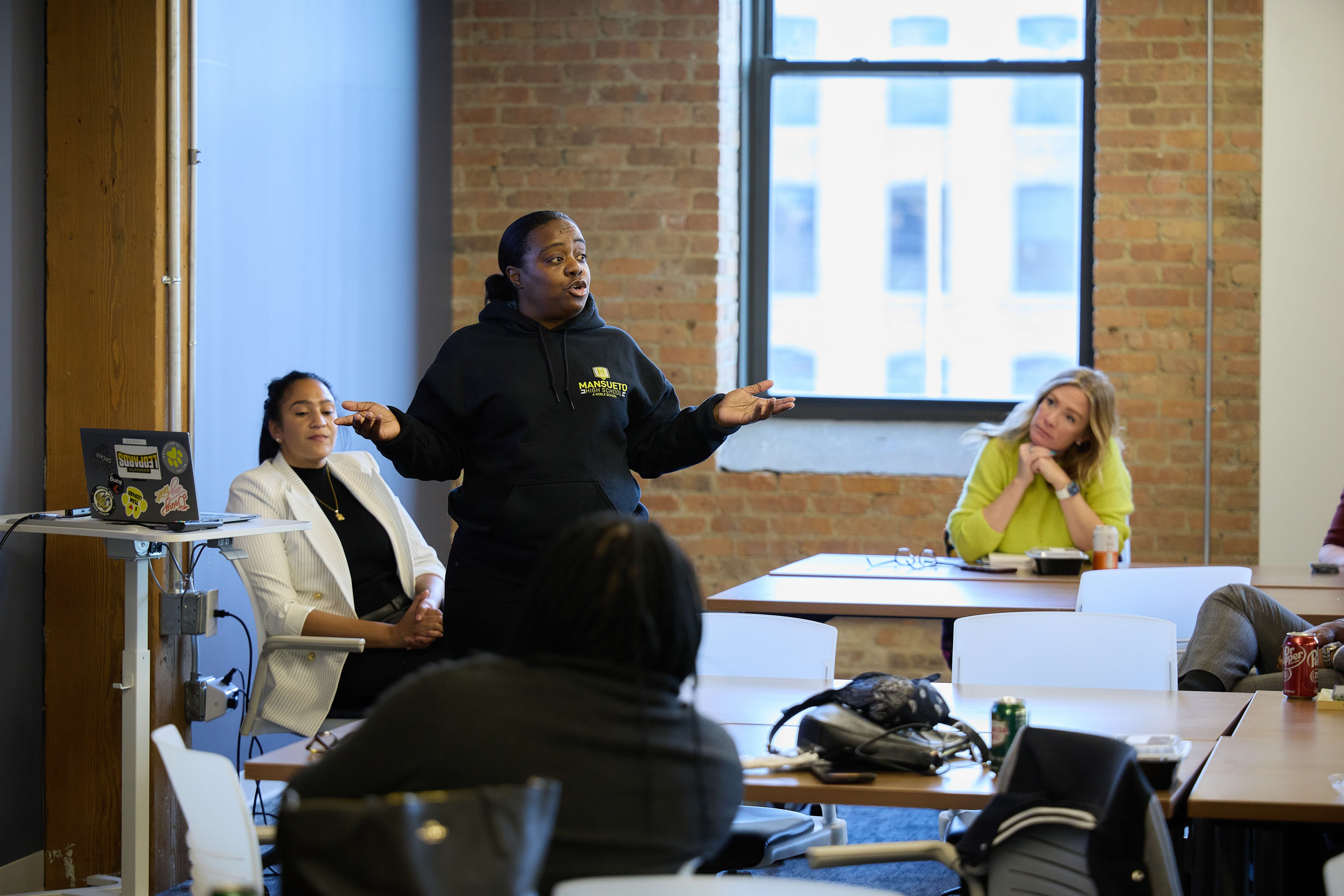 Myisha Shields, a parent at Mansueto College Prep, speaks to a crowds, both her hands up in a shrugging kind of gesture. She is a Black woman with straight black hair pulled back into a short ponytail and brown skin. She is wearing a black hoodie with the bright yellow and gray Mansueto logo on the breast pocket area.