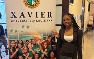 A Rowe-Clark Math and Science Academy student stands in front of a sign for Xavier University of Louisiana, a historically Black university in New Orleans. She is wearing a business casual outfit and throwing up a peace sign.