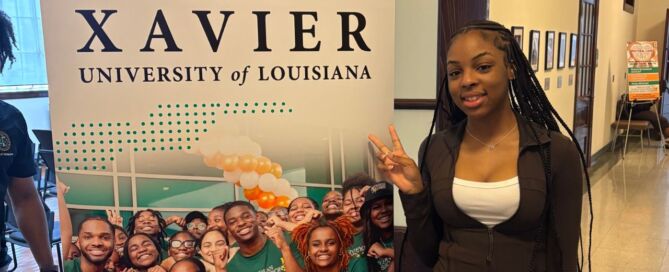 A Rowe-Clark Math and Science Academy student stands in front of a sign for Xavier University of Louisiana, a historically Black university in New Orleans. She is wearing a business casual outfit and throwing up a peace sign.