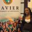 A Rowe-Clark Math and Science Academy student stands in front of a sign for Xavier University of Louisiana, a historically Black university in New Orleans. She is wearing a business casual outfit and throwing up a peace sign.