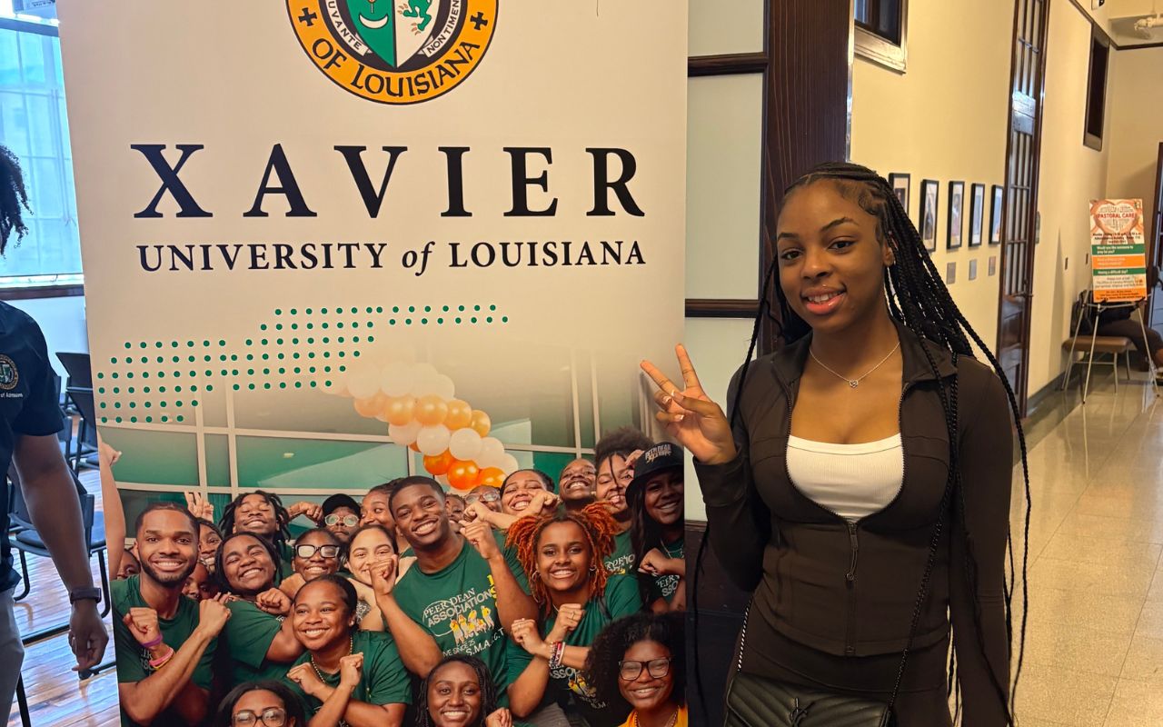 A Rowe-Clark Math and Science Academy student stands in front of a sign for Xavier University of Louisiana, a historically Black university in New Orleans. She is wearing a business casual outfit and throwing up a peace sign.