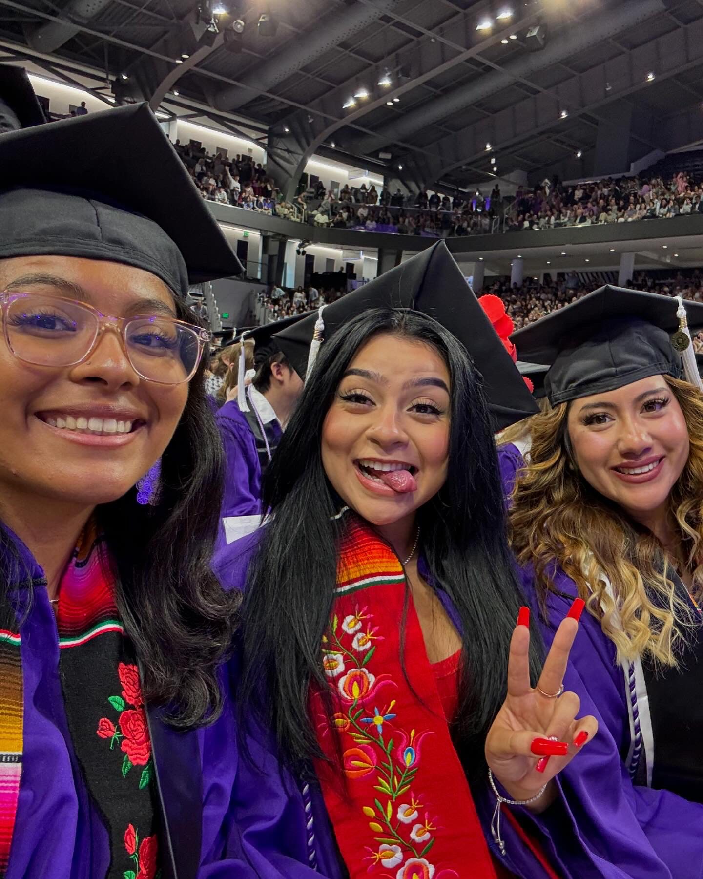 aileen at college graduation Galeana at her college graduation. She is wearing a black graduation cap and purple graduation robes with a bright red sash, embroidered with flowers and many other colors. She sits in between two friends as they take a selfie. She is throwing up a peace sign with gorgeous long matching red nails and sticking her tongue out humorously. Galeana is a Latine woman with long straight black hair and light brown skin.
