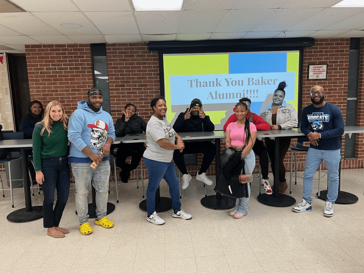 Dixon and Justin Harris stand with a group of alumni and other staff around a long table in a Baker common space. Behind them, you can see a screen with the words "Thank you, Baker Alumni!!!" projected onto it.