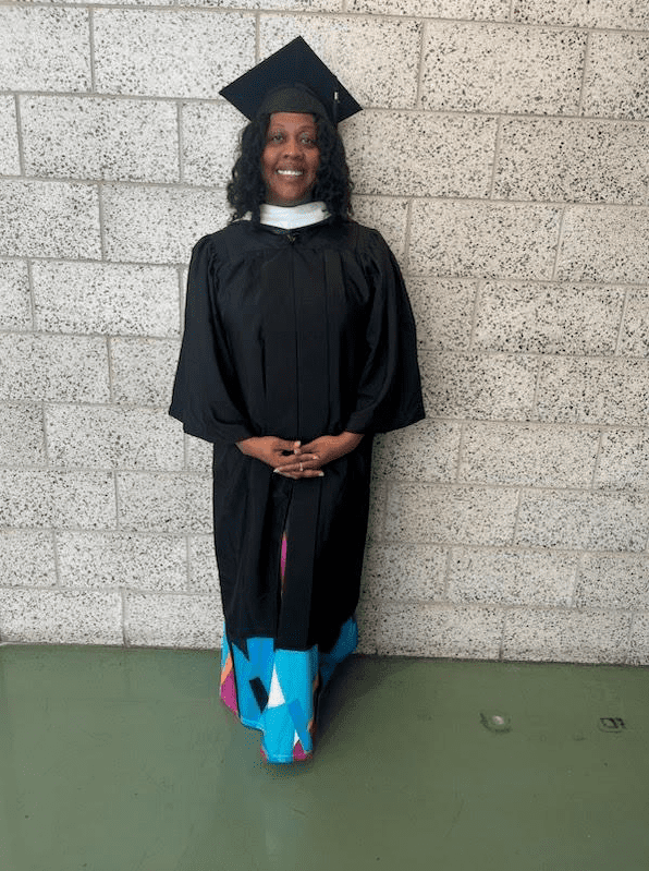 Selena Dixon stands against a gray brick wall, smiling and posing for a photo in black graduation robes over a colorful dress. She is a Black woman with black wavy hair at shoulder-length and brown skin.