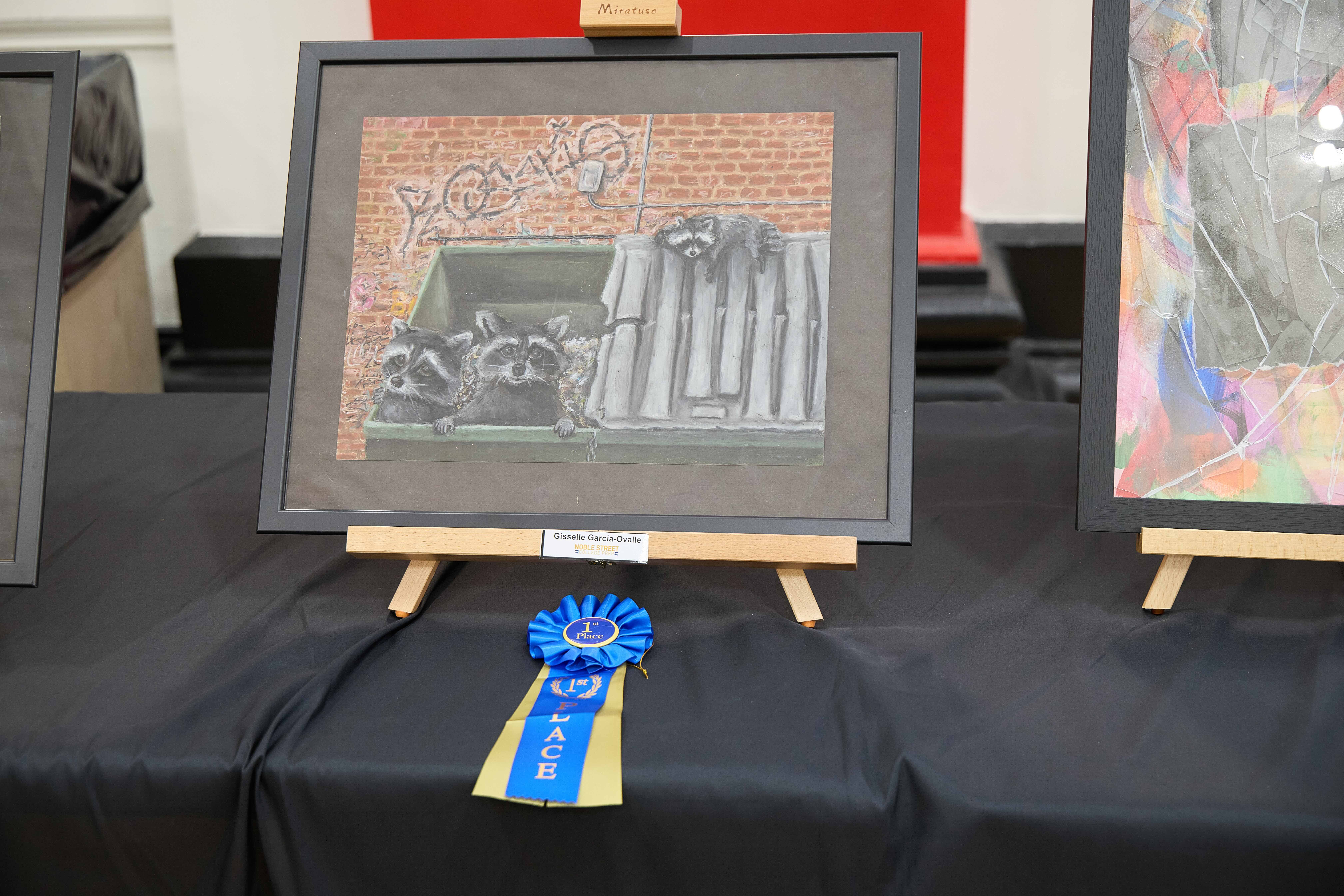 2D Design 1st Place A drawing of raccoons in a dumpster sits on a small easel on a table. You can see a bright blue 1st place ribbon in front of it. Two raccoons are peeking out from inside the dumpster, one looks at the camera. Another raccoon is dozing on the dumpster lid that covers the other half of the container. Behind the dumpster, there is a red. brick wall with graffiti on it.