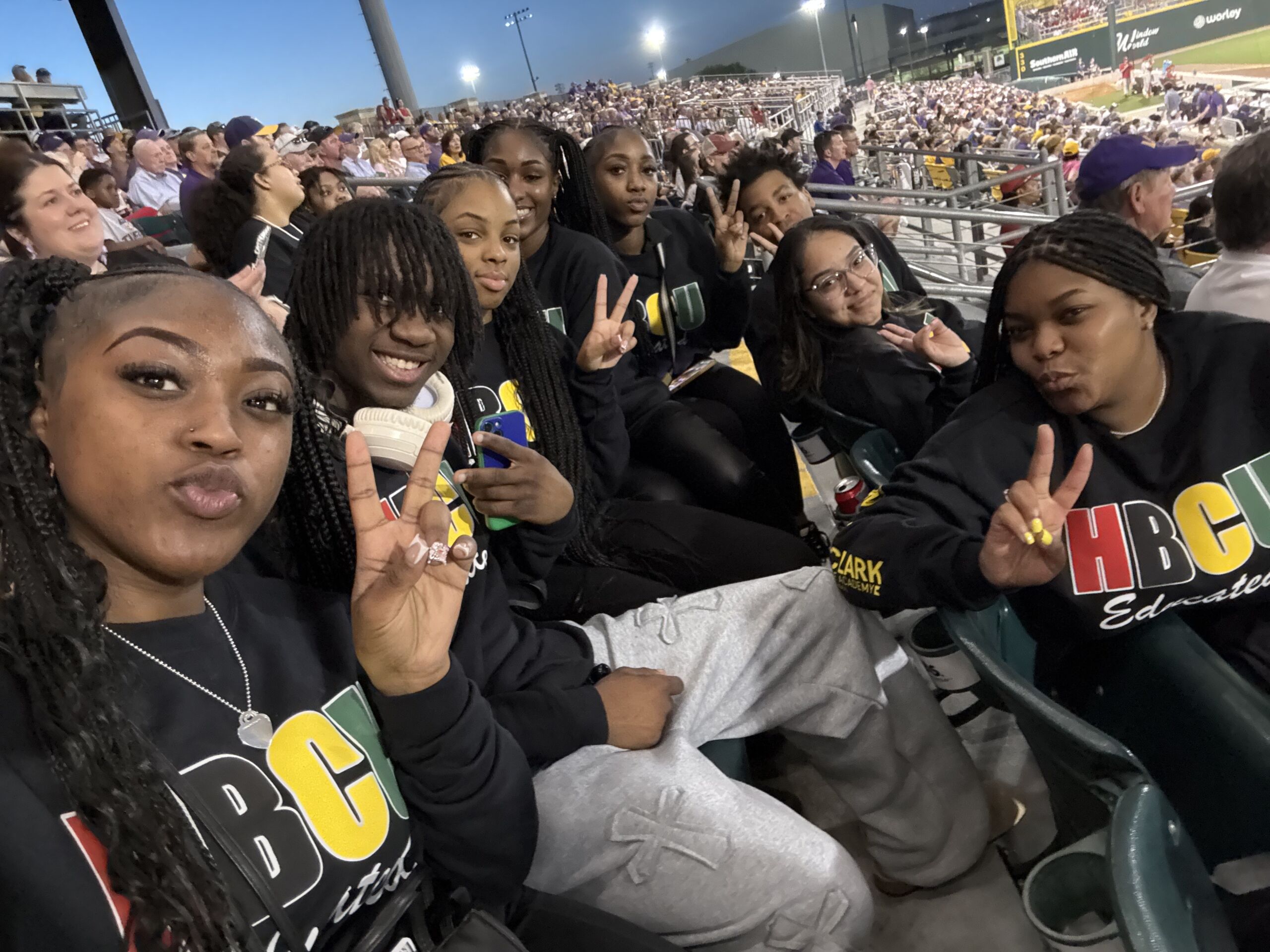 Rowe-Clark students take a selfie in the stands of the baseball stadium. Many of them are throwing up peace signs and wearing black sweatshirts that say HBCU on them in blocky letters in the pan-African colors: red, black, yellow, and green