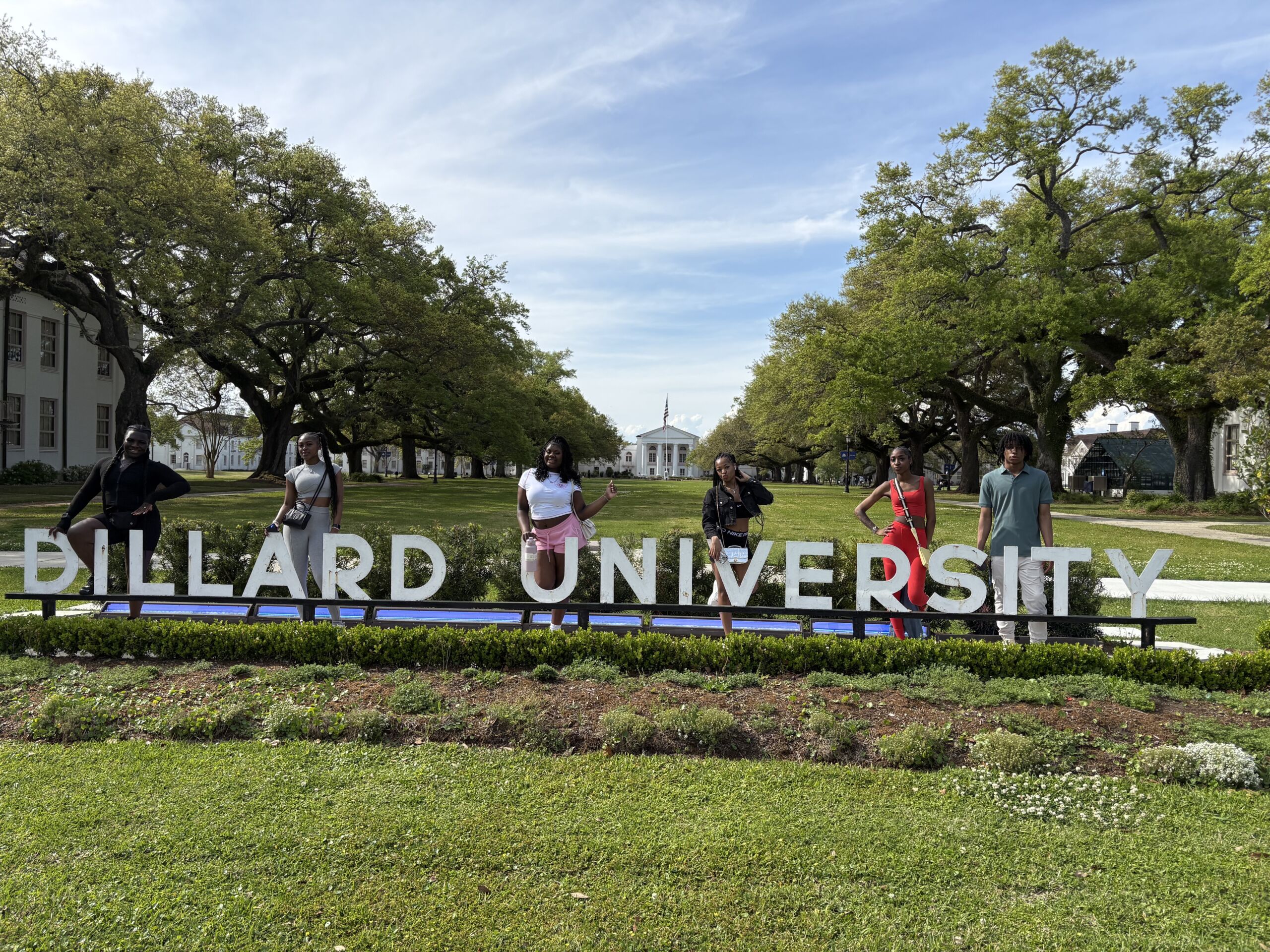 Six Rowe-Clark students stand behind the Dillard University sign in a line. It's a bright sunny day and you can see the trees, fields, and buildings of the campus behind them.