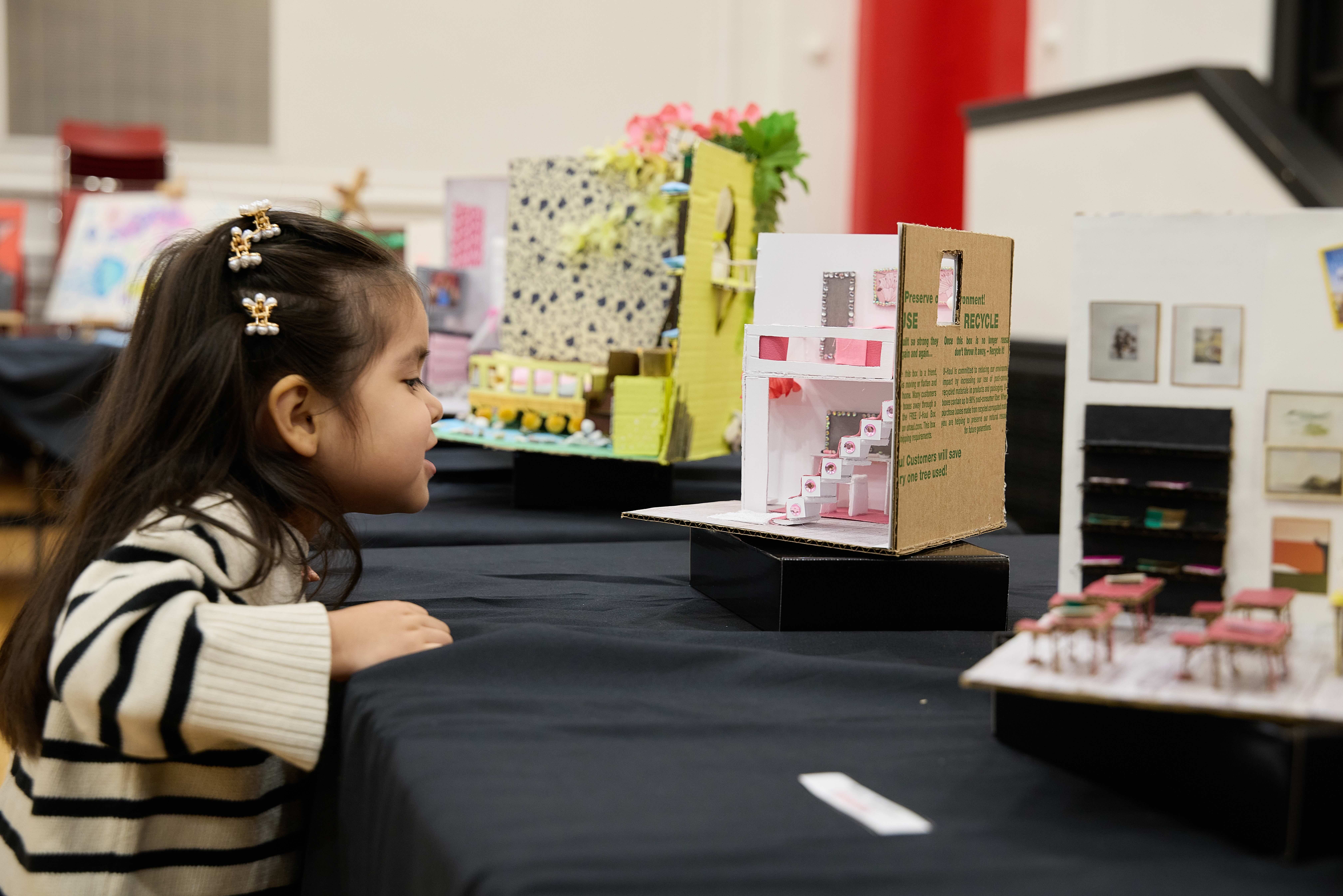 A young girl leans on a table to get a closer look at a cardboard diorama of a pink and white bedroom.