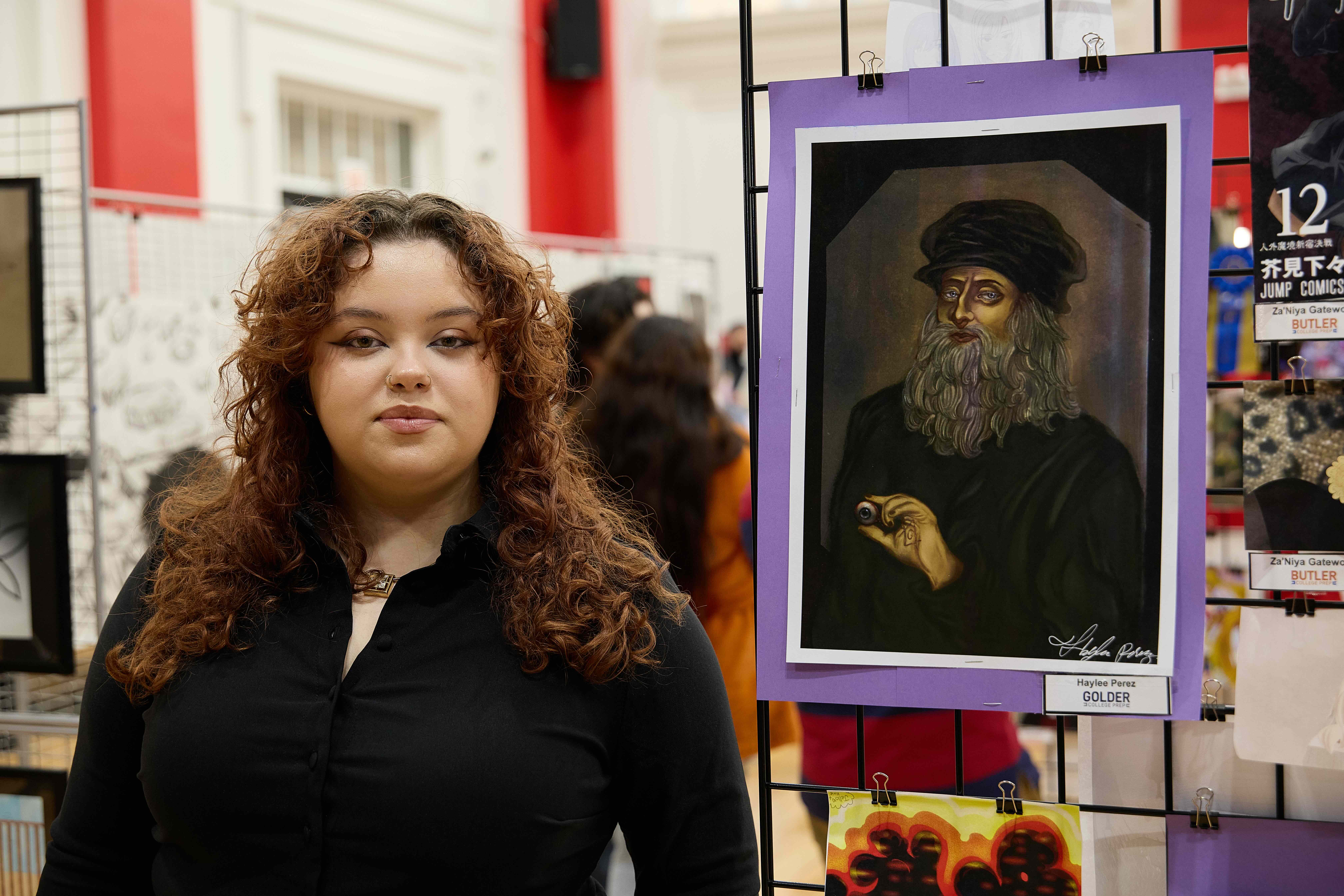 Haylee Perez, a student at Golder College Prep, stands next to her digital painting. The subject is an older man with a long gray beard and wearing flowing black robes and a black soft hat. He is holding an eyeball in between two fingers and has a sigil etched into the palm of his hand that has the letter "G" in the center of it. He is framed by a black vignette border with a nondescript brown and gray background behind him. Haylee wears a matching black button-up and a short necklace with a square gold and crystal pendant. She has long red curly hair and brown skin.
