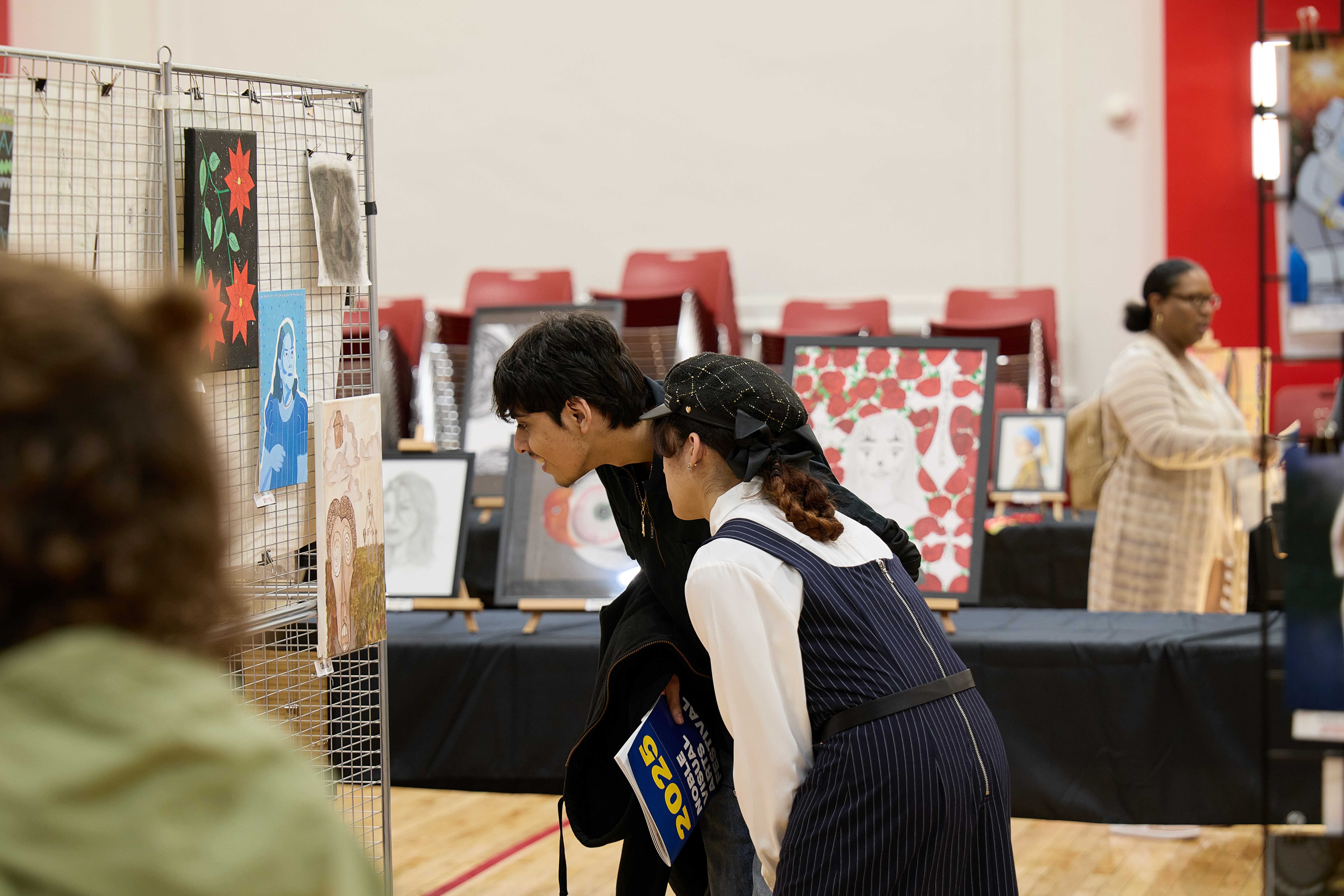 Two students lean in to view a painting hung up on a rack. It is sepia-toned and shows an abstract face of a person screaming in front of a field with tall grasses with a skeleton standing in it. Looming above the face is a small house that sits in the middle of clouds in the sky.