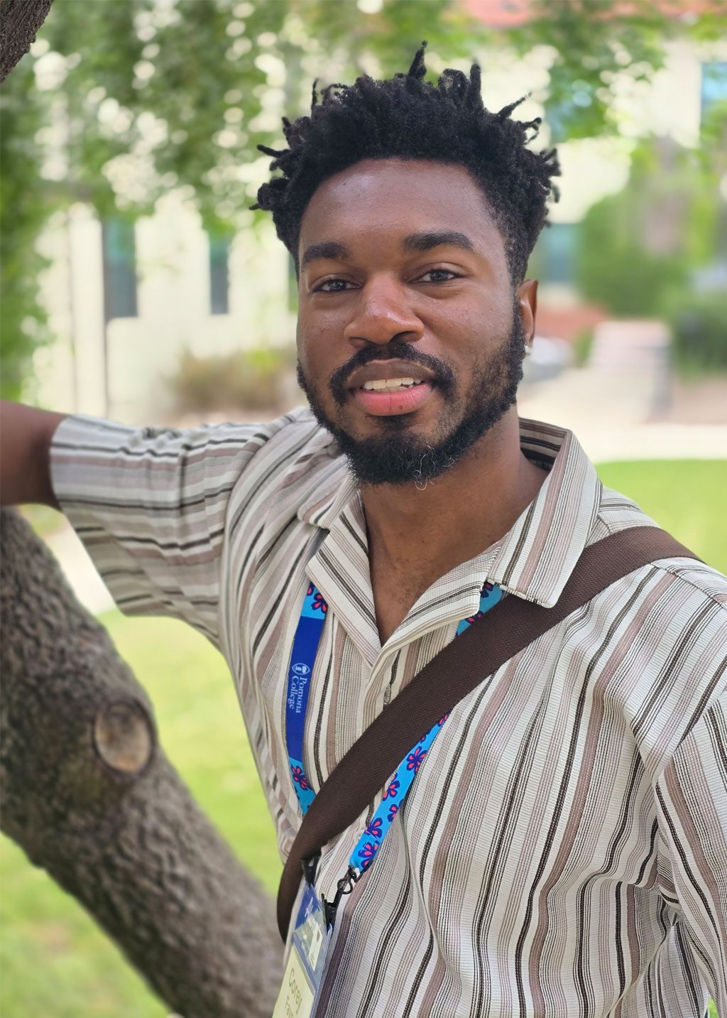 Corey Fayne DLF Cohort 2025 Headshot of Corey Fayne. He is a Black man with a beard and short black spike locs. He is smiling standing next to a tree, and wearing a brown colored striped shirt.