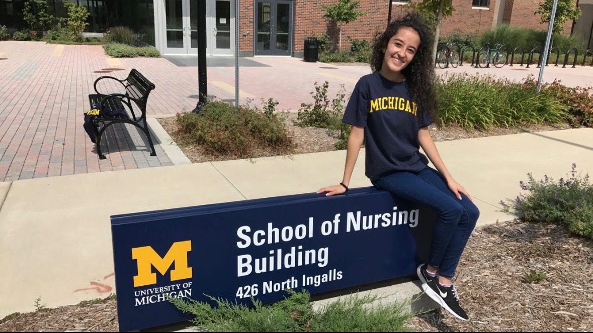 Screenshot Jai sits on the school sign for University of Michigan's School of Nursing Building. Jai is smiling brightly, her head tilted as she looks at the camera. A bright yellow "Michigan" is emblazoned on her dark blue t-shirt.