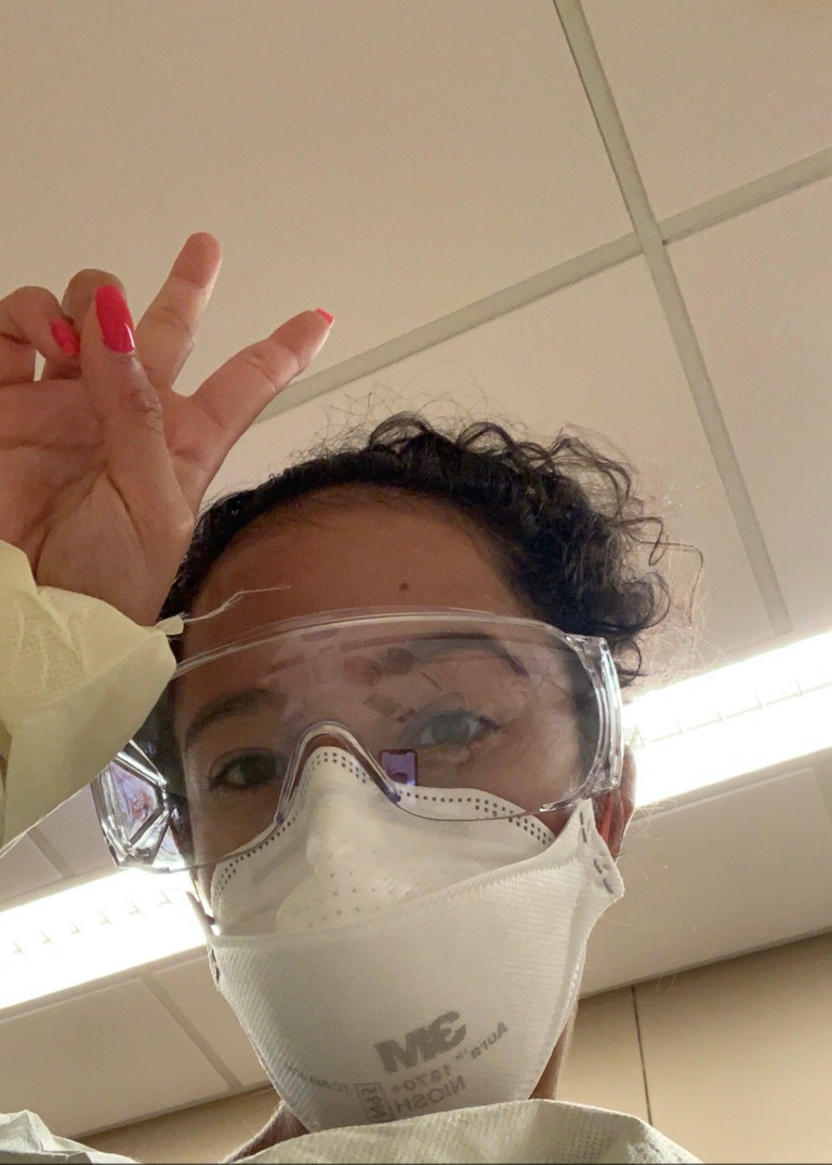 Screenshot Jai takes a selfie of her in a white medical mask and goggles. She's holding up a peace sign. You can see the white and gray panelled ceiling and fluorescent lights of the doctor's office above her.