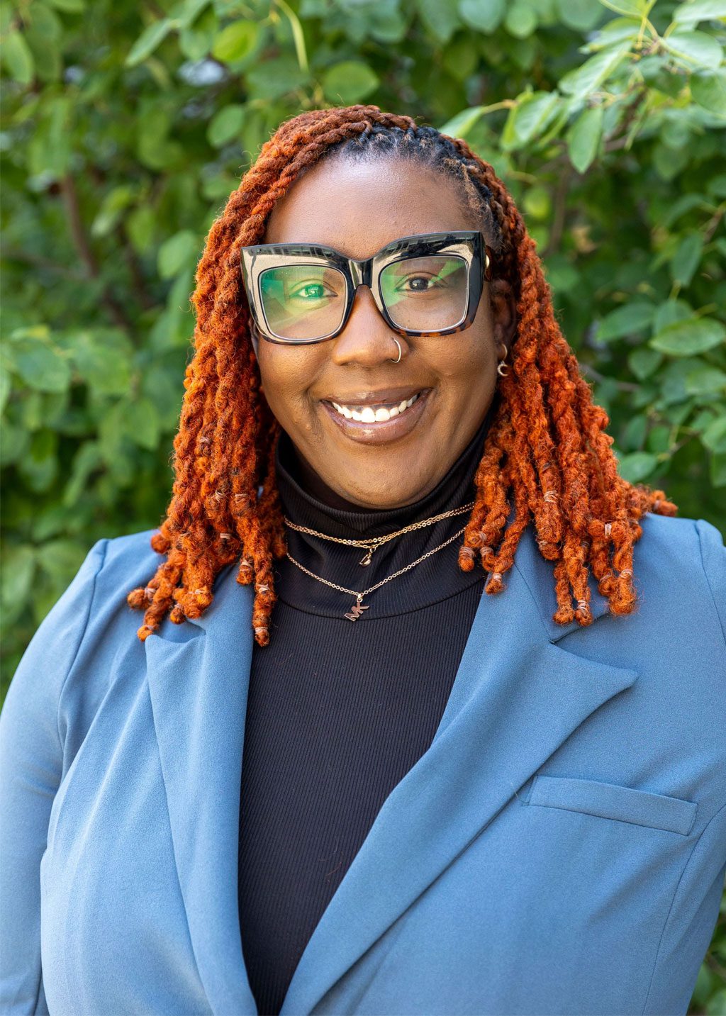 Maisa Reid DLF Cohort 2025 Headshot of Maisa Reid. She is a Black woman and short black and orange locs that go down to her shoulders. She is smiling and wearing thick framed glasses and a black turtle neck shirt and blue blazer.