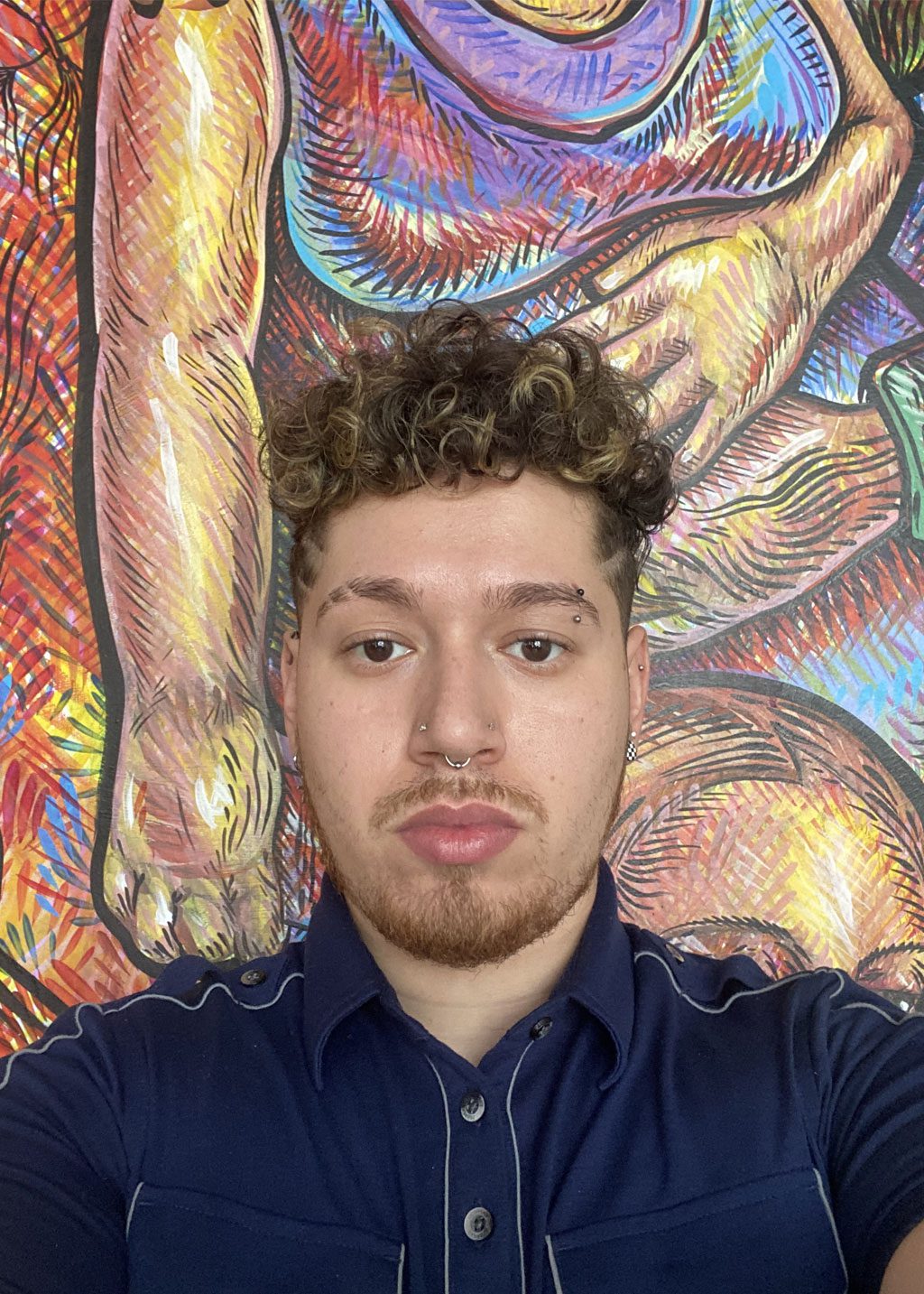 Roy Jaime DLF Cohort 2025 Headshot of Roy Jaime. He is a Hispanic man with curly light brown hair, highlights and light beard. He has piercings in his left eye brow, right nostril and nose ring. Wearing a navy blue shirt with white thin stripes on the shoulders, and front pockets.