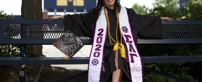 Jailin Morales, an alum of Noble Street College Prep in Chicago, IL, sits on a bench in graduation robes. She is smiling brightly while a big University of Michigan "M" sign sits in the distance over her shoulder. Her graduation cap sits beside her and is decorated with a bedazzled syringe and stethoscope with the words "time for shots". She is a young Latine woman with medium length curly black hair.