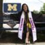 Jailin Morales, an alum of Noble Street College Prep in Chicago, IL, sits on a bench in graduation robes. She is smiling brightly while a big University of Michigan "M" sign sits in the distance over her shoulder. Her graduation cap sits beside her and is decorated with a bedazzled syringe and stethoscope with the words "time for shots". She is a young Latine woman with medium length curly black hair.