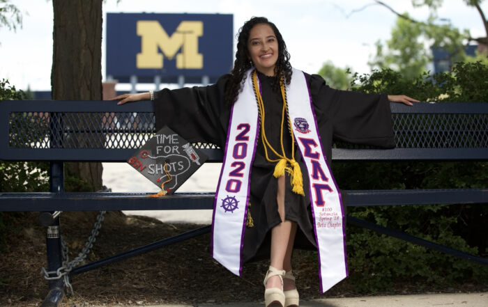 Jailin Morales, an alum of Noble Street College Prep in Chicago, IL, sits on a bench in graduation robes. She is smiling brightly while a big University of Michigan "M" sign sits in the distance over her shoulder. Her graduation cap sits beside her and is decorated with a bedazzled syringe and stethoscope with the words "time for shots". She is a young Latine woman with medium length curly black hair.