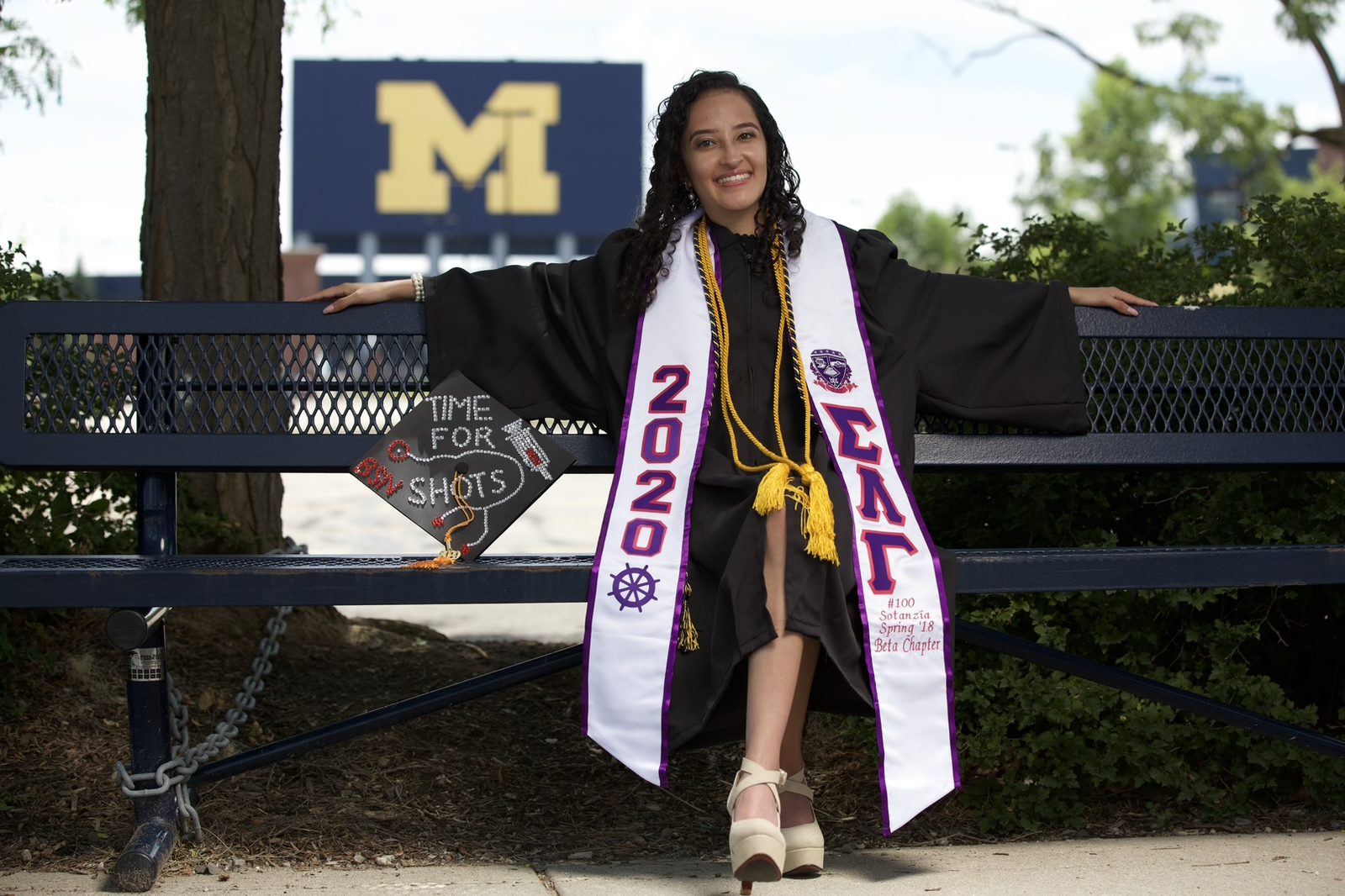 Jailin Morales, an alum of Noble Street College Prep in Chicago, IL, sits on a bench in graduation robes. She is smiling brightly while a big University of Michigan "M" sign sits in the distance over her shoulder. Her graduation cap sits beside her and is decorated with a bedazzled syringe and stethoscope with the words "time for shots". She is a young Latine woman with medium length curly black hair.