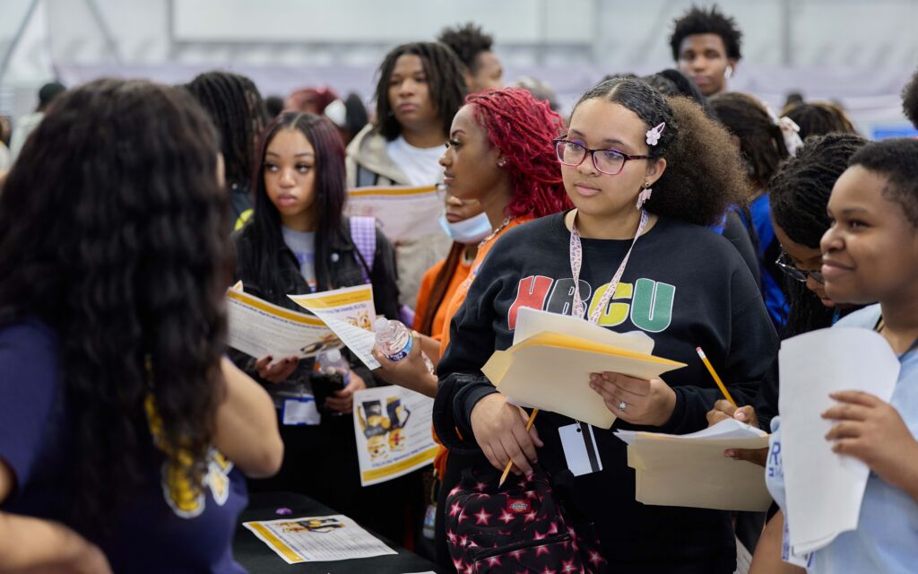 A student looks in rapt attention to an HBCU admissions counselor at Noble Schools' 2025 HBCU College Fair. She's wearing a black sweatshirt that has "HBCU" in the pan-African colors blazoned across the front.