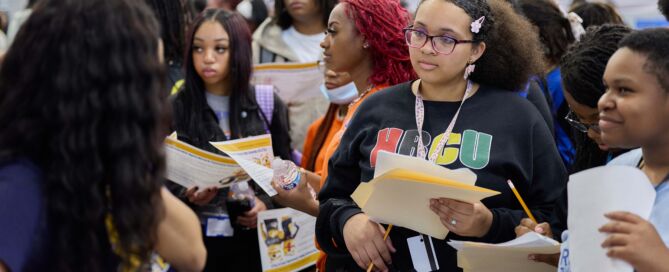 A student looks in rapt attention to an HBCU admissions counselor at Noble Schools' 2025 HBCU College Fair. She's wearing a black sweatshirt that has "HBCU" in the pan-African colors blazoned across the front.