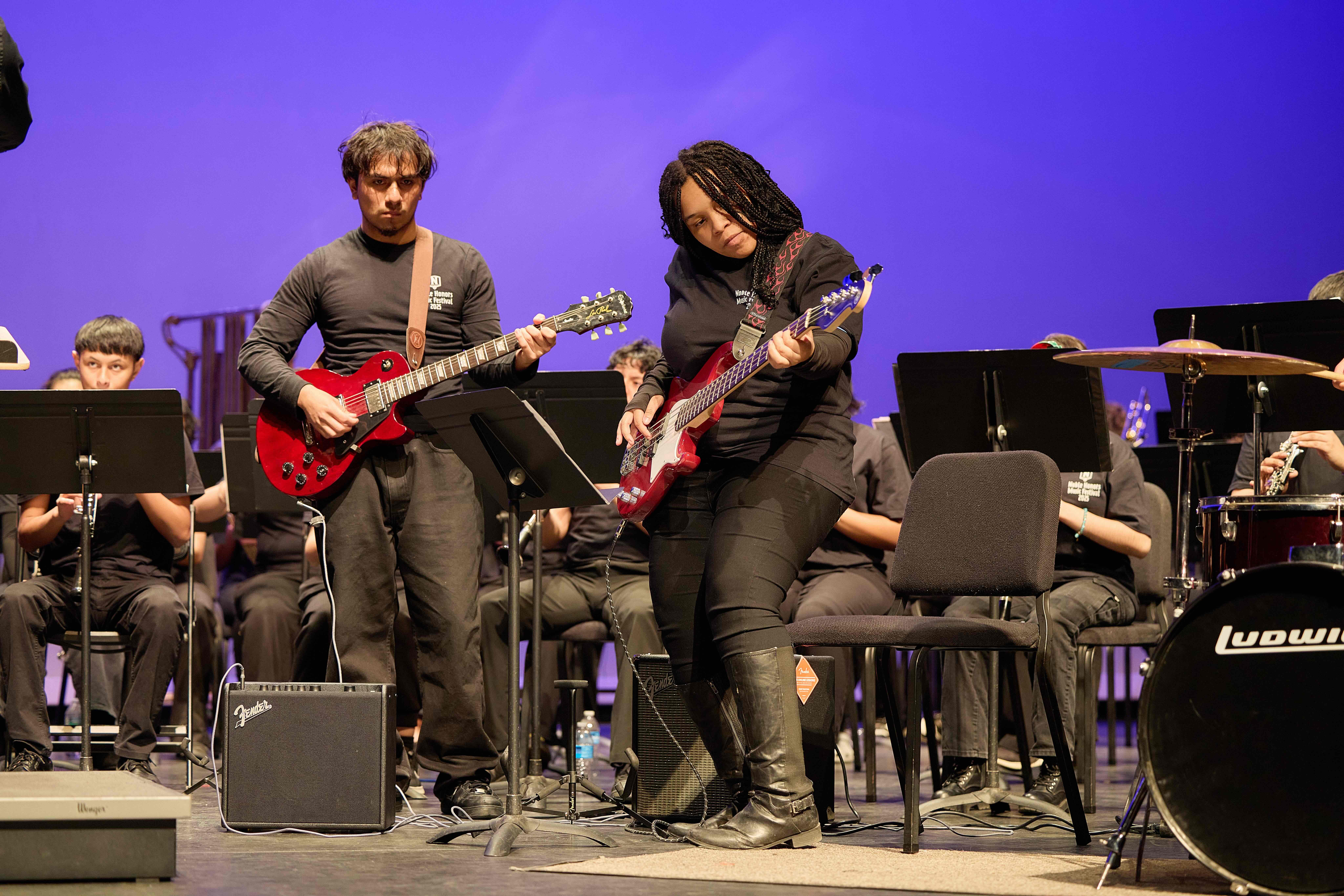 jacob playing guitar Jacob plays his bright red electric guitar on stage. He is standing next to his peer who is playing bass. Behind him, you can see the rest of the ensemble and a bright indigo backdrop. He looks very focused, staring straight ahead. Both Jacob and the rest of the students are wearing all black with a matching t-shirt that has a white Noble Schools logo on the upper right corner with the words "Noble Honors Music Festival 2025" underneath it.
