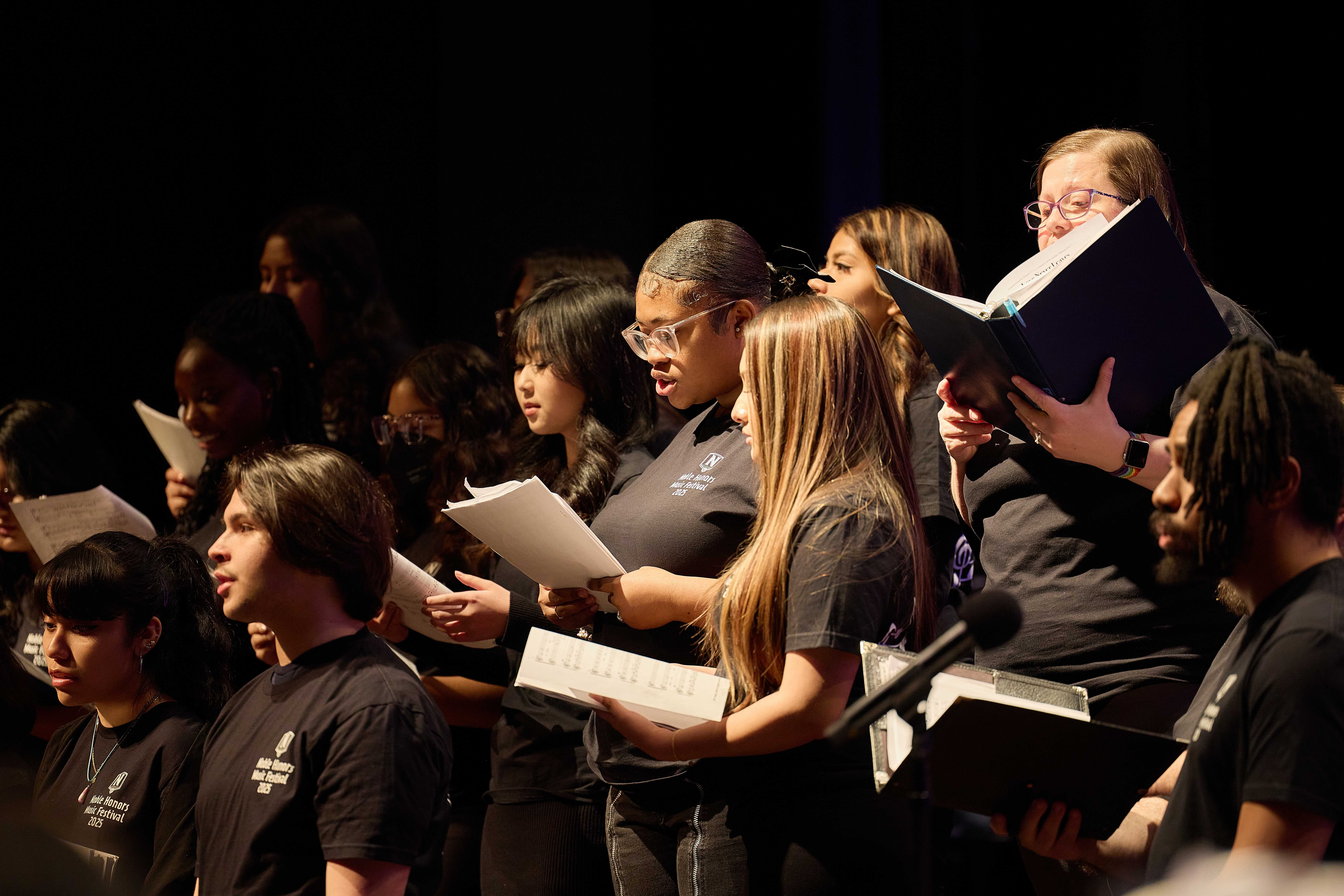 kemariya and lyndia in choir Lyndia and Kemariya stand side-by-side and sing in the middle of their choir ensemble. They're reading from sheet music in their hands.