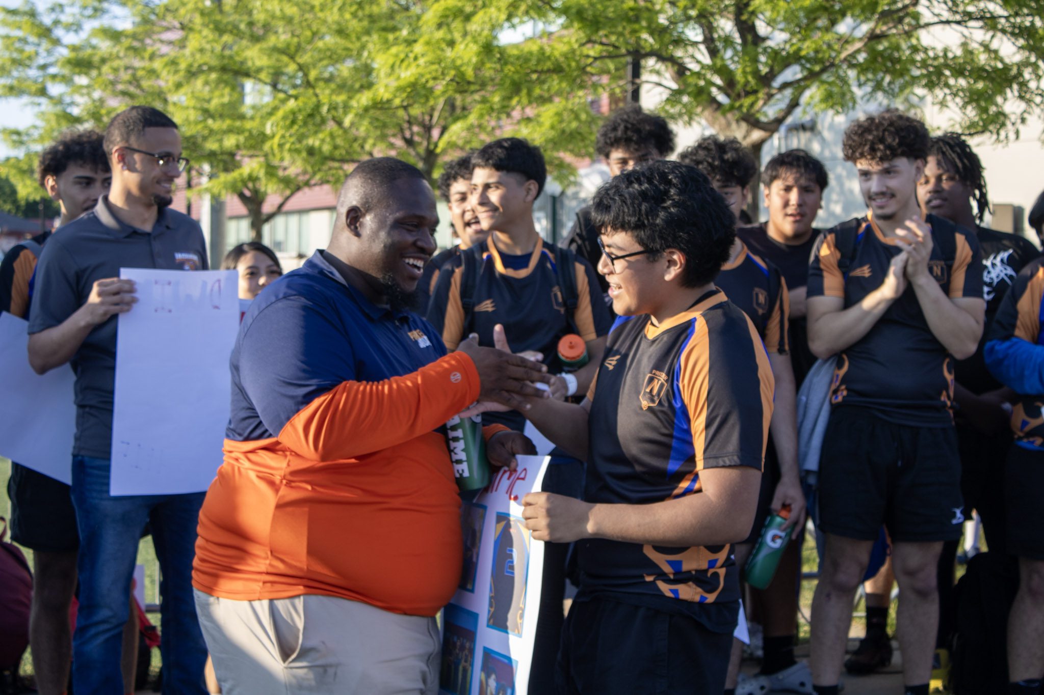 Johnson Coaching Johnson and a student meet each other with a dap, both smiling widely. The student is holding a poster with his photos in his other hand. The rest of the Pritzker College Prep rugby team claps around them.