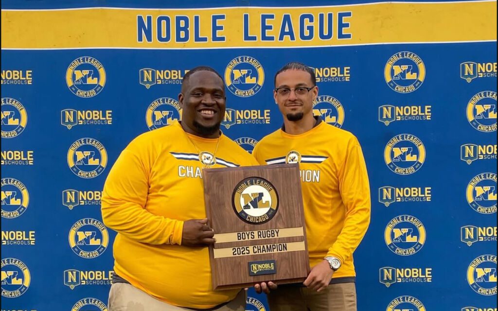 Pritzker College Prep coaches Jovon Hernandez and Anthony Johnson pose for a photo, holding a plaque between them. The plaque says “Boys Rugby 2025 Champions”.