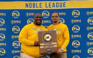Pritzker College Prep coaches Jovon Hernandez and Anthony Johnson pose for a photo, holding a plaque between them. The plaque says “Boys Rugby 2025 Champions”.