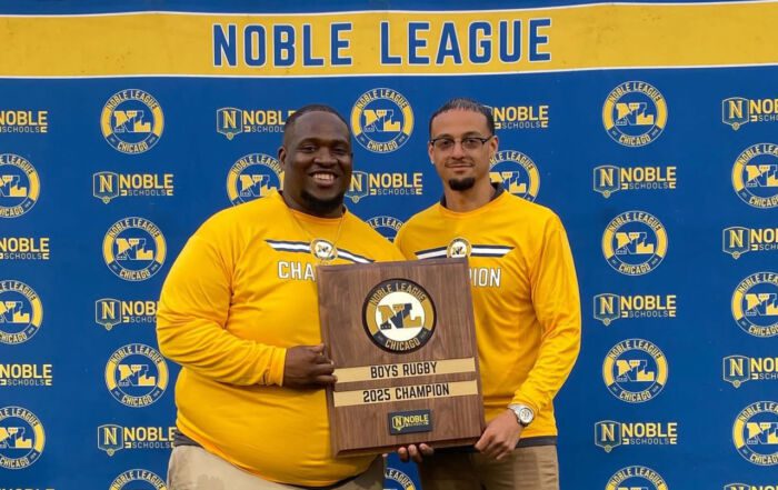 Pritzker College Prep coaches Jovon Hernandez and Anthony Johnson pose for a photo, holding a plaque between them. The plaque says “Boys Rugby 2025 Champions”.
