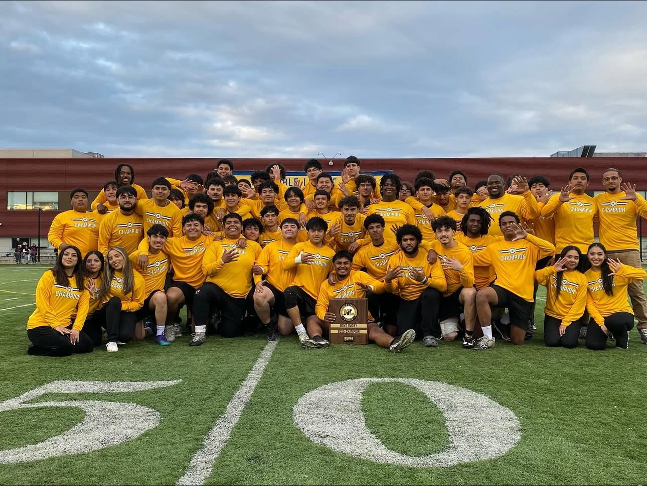 Pritzker Rugby Team SY25 Photo The whole Pritzker College Prep rugby team poses for a photo in their bright yellow jerseys at the 50-yard line on the field. The student sitting in the middle holds a plaque that says “Boys Rugby 2025 Champions.”