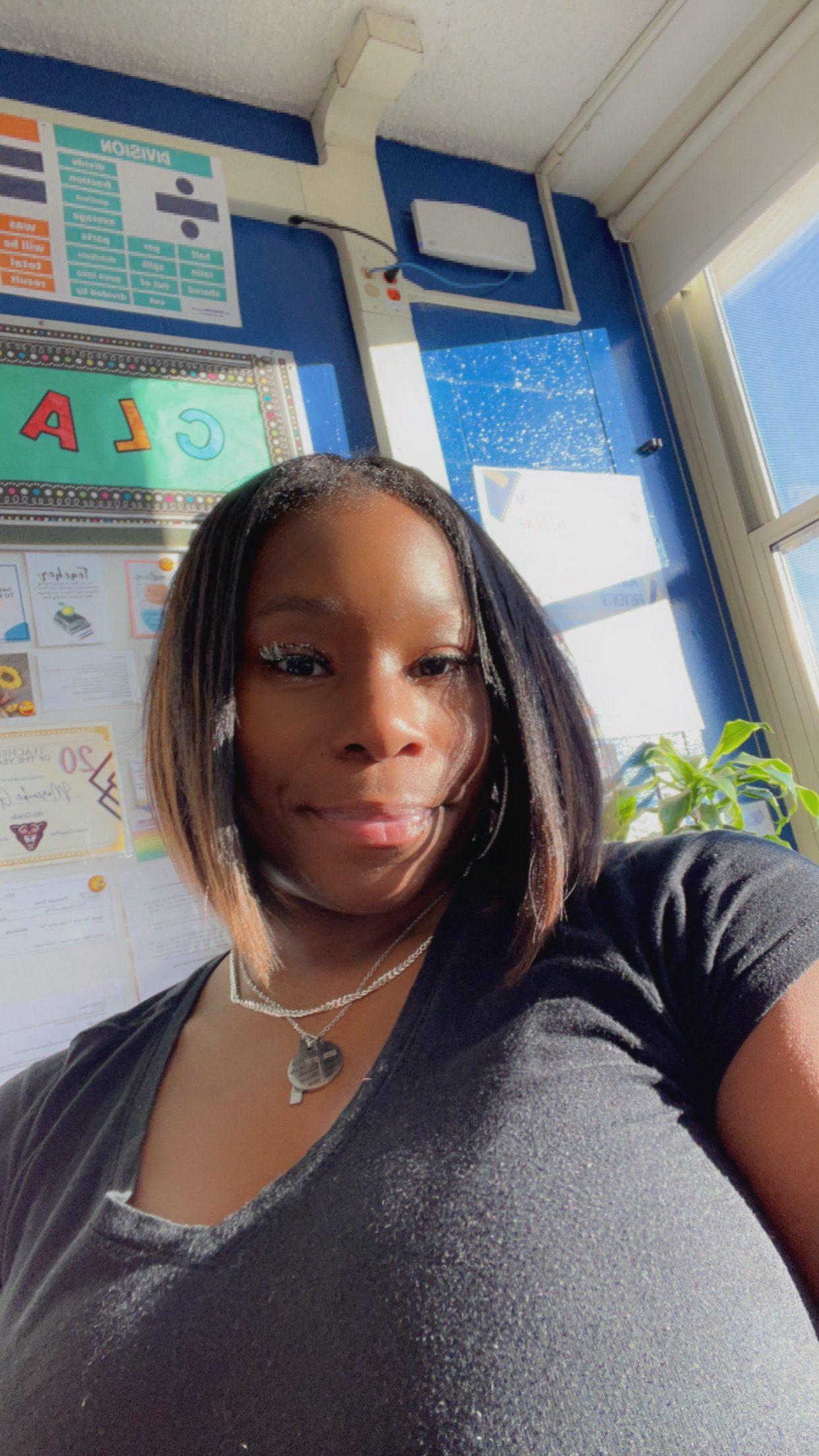 A headshot of Morenike Giwa. She is a Black woman and is wearing a short sleeve black blouse. You can see her classroom board behind her.
