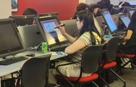 A Noble student in a college classroom sits at a giant tablet, using a pen to draw on it.