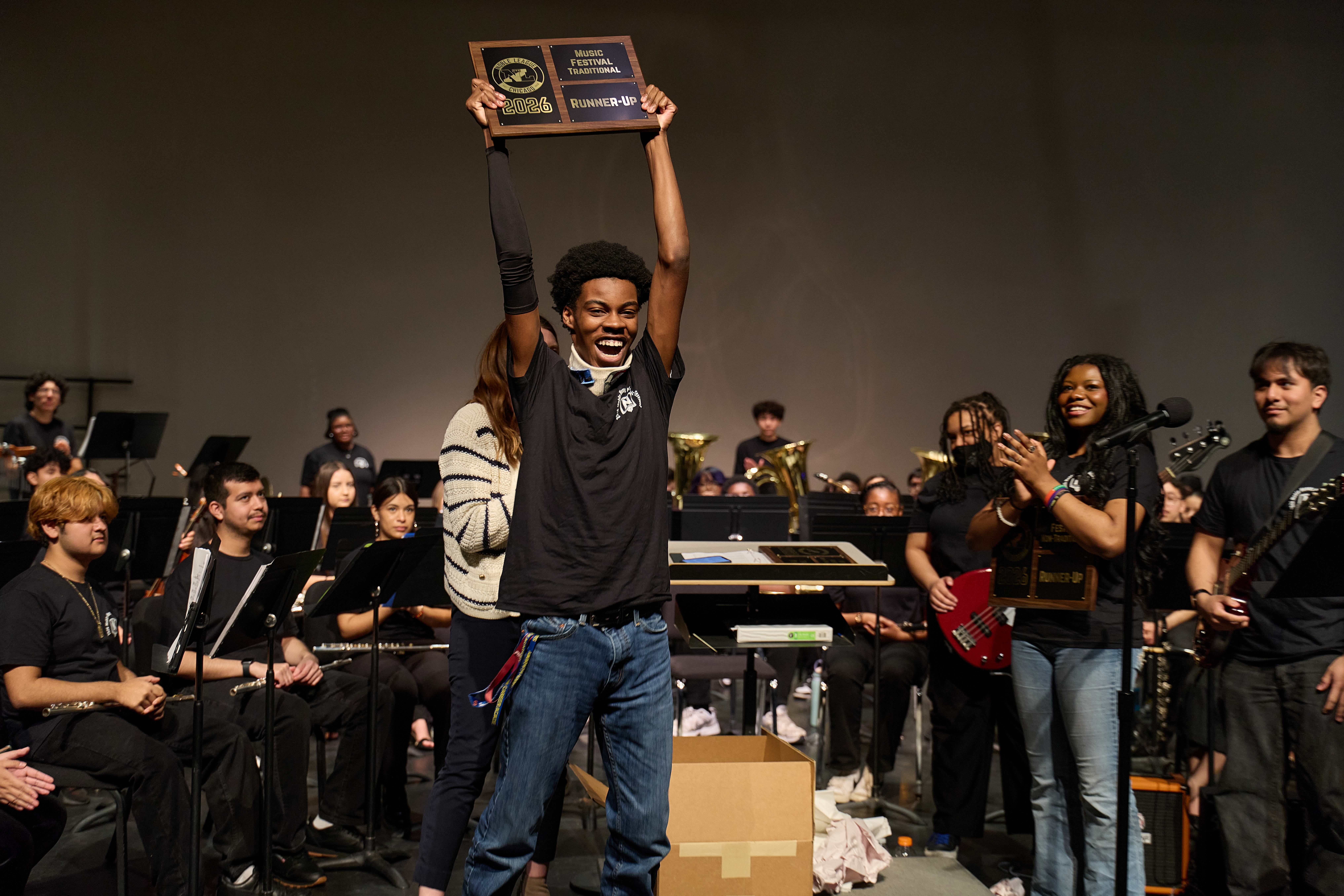 A student holds up a plaque, celebrating, on the stage at the Music Honors Festival. Musicians in the ensemble clap behind him. The plaque says "Runner-Up, Music Festival, Traditional"