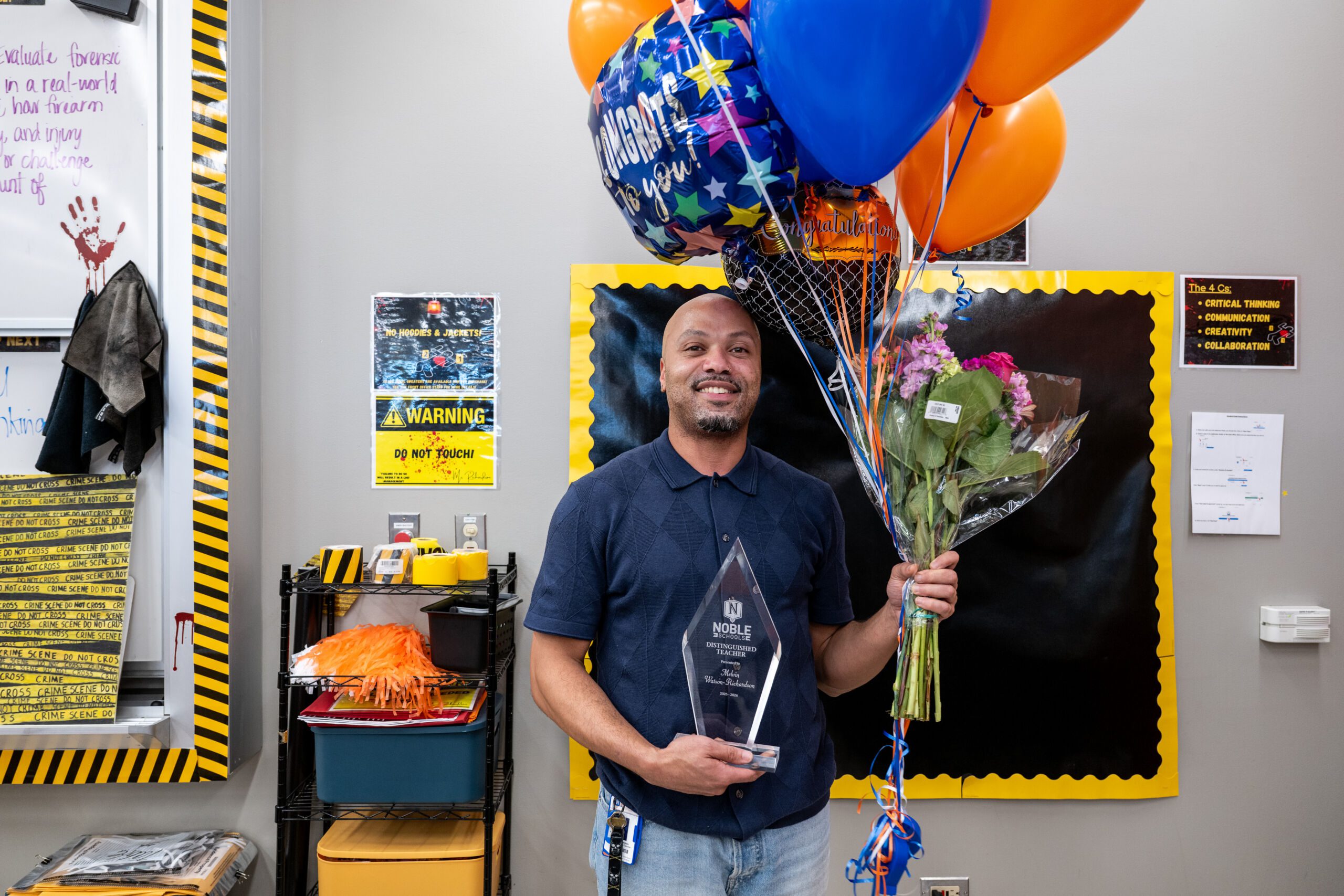 CD2_1961 Melvin Watson-Richards smiles, holding his glass Distinguished Teacher plaque in one hand and balloons in the other. He is standing in his classroom.