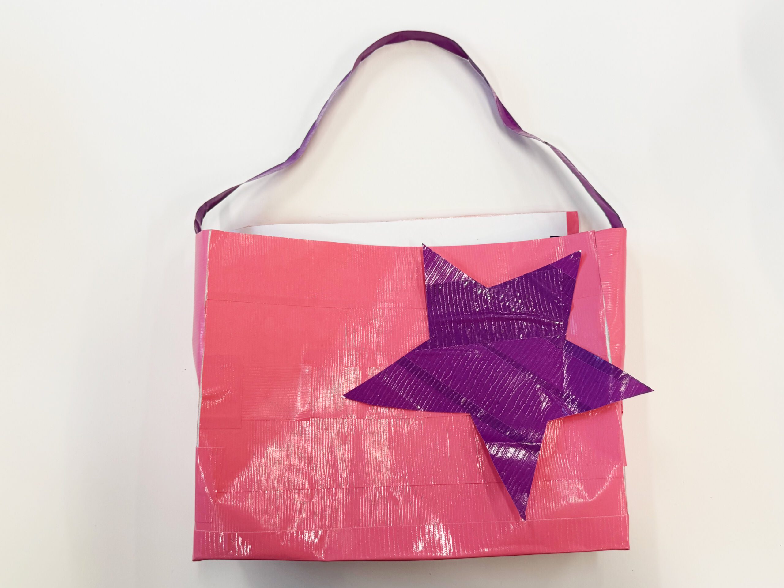 A small hand purse constructed with duck tape. The main square body of the purse is a bright rose punk and a big purple star is stuck on the front of it. The thin strap at the top is the same purple.