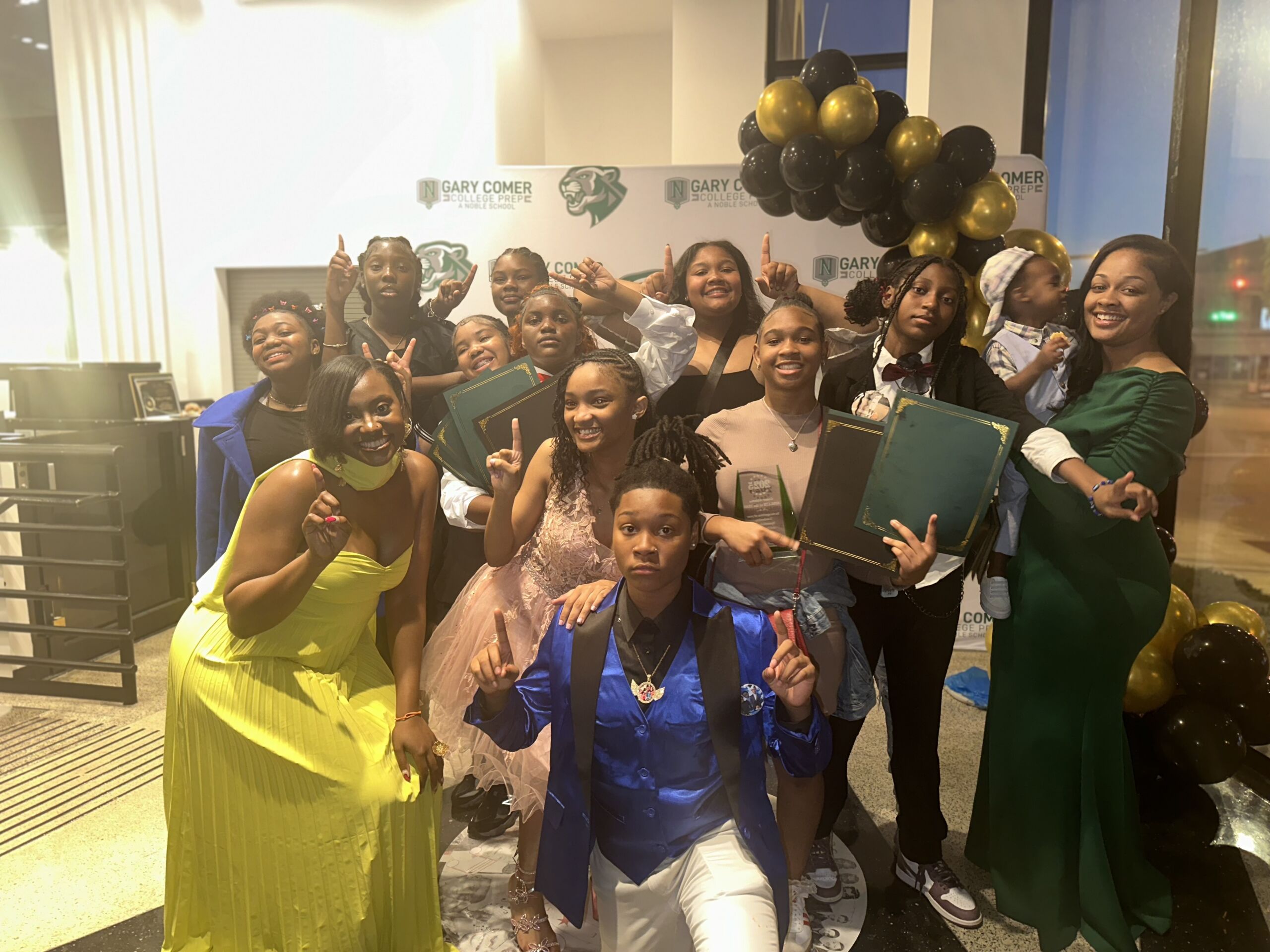 Myers and her team of students are dressed up and posing for a photo in front of a Gary Comer branded backdrop. Some of them hold dark green award certificate folders in their hands.