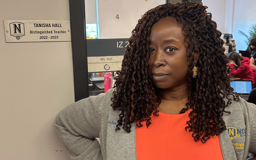 Ta-Tanisha Hall, an African American Literature teacher at ITW David Speer Academy in Chicago, IL, stands at the door to her classroom. She is smiling confidently and standing next to the plaque on the side of her door that says "Tanisha Hall, Distinguished Teacher, 2022-2023"