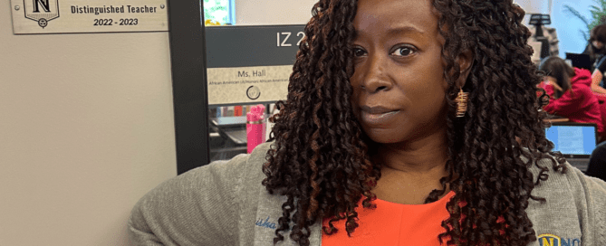 Ta-Tanisha Hall, an African American Literature teacher at ITW David Speer Academy in Chicago, IL, stands at the door to her classroom. She is smiling confidently and standing next to the plaque on the side of her door that says "Tanisha Hall, Distinguished Teacher, 2022-2023"