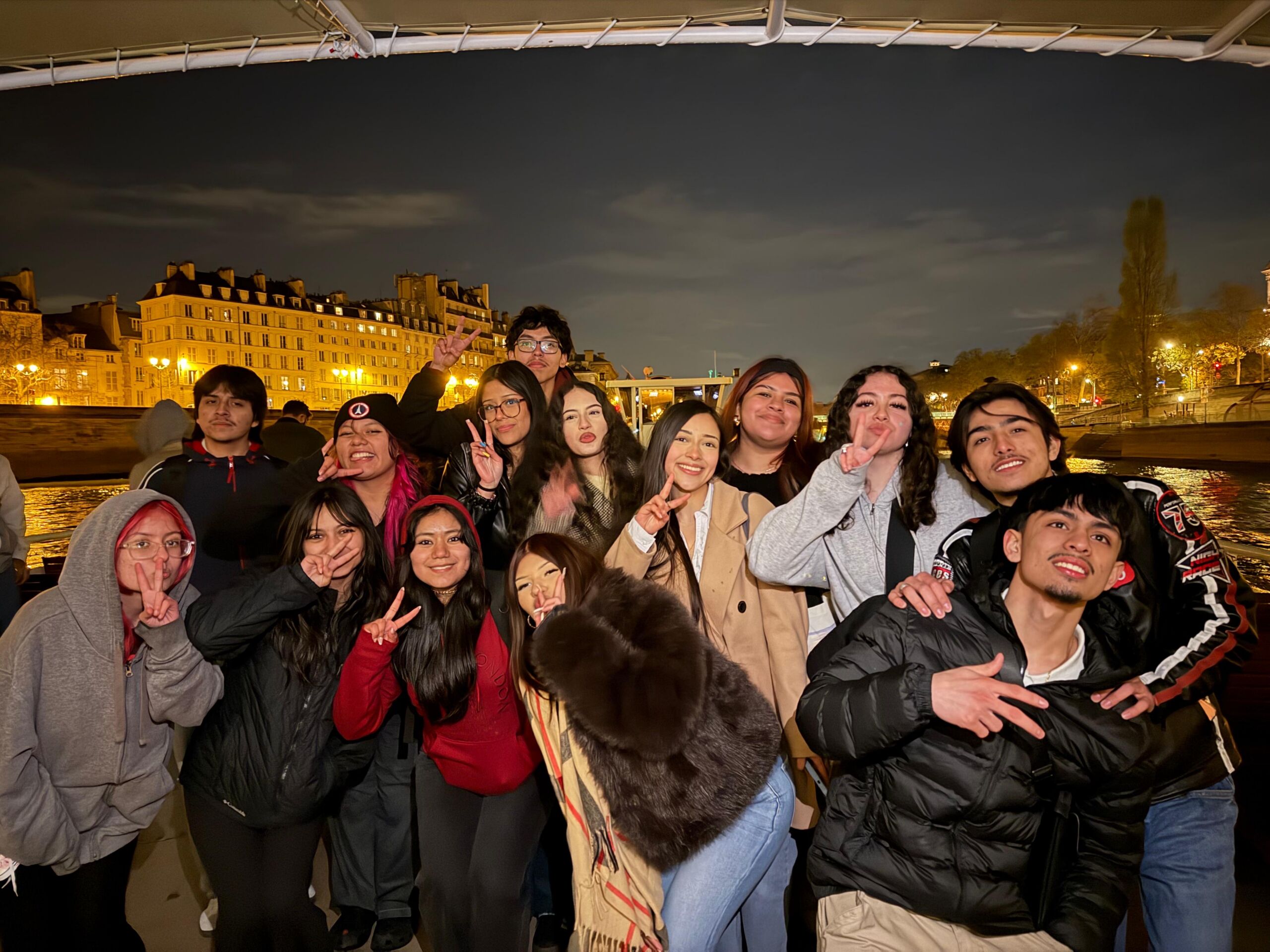 untitled (3) A group of students and teachers gather for a photo on the boat. You can see the Seine River and the city buildings of Paris lit up behind them.