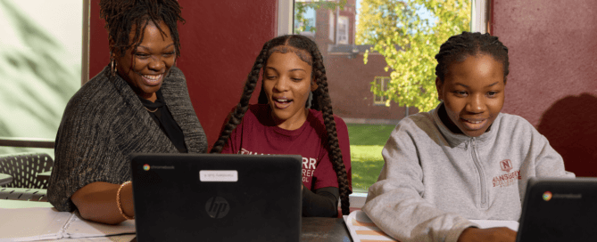 A teacher at Hansberry College Prep stands behind a student, helping her with something on her laptop. They are both smiling and talking. Another student sits nearby, also focused on her laptop.