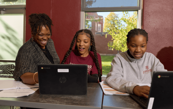 A teacher at Hansberry College Prep stands behind a student, helping her with something on her laptop. They are both smiling and talking. Another student sits nearby, also focused on her laptop.