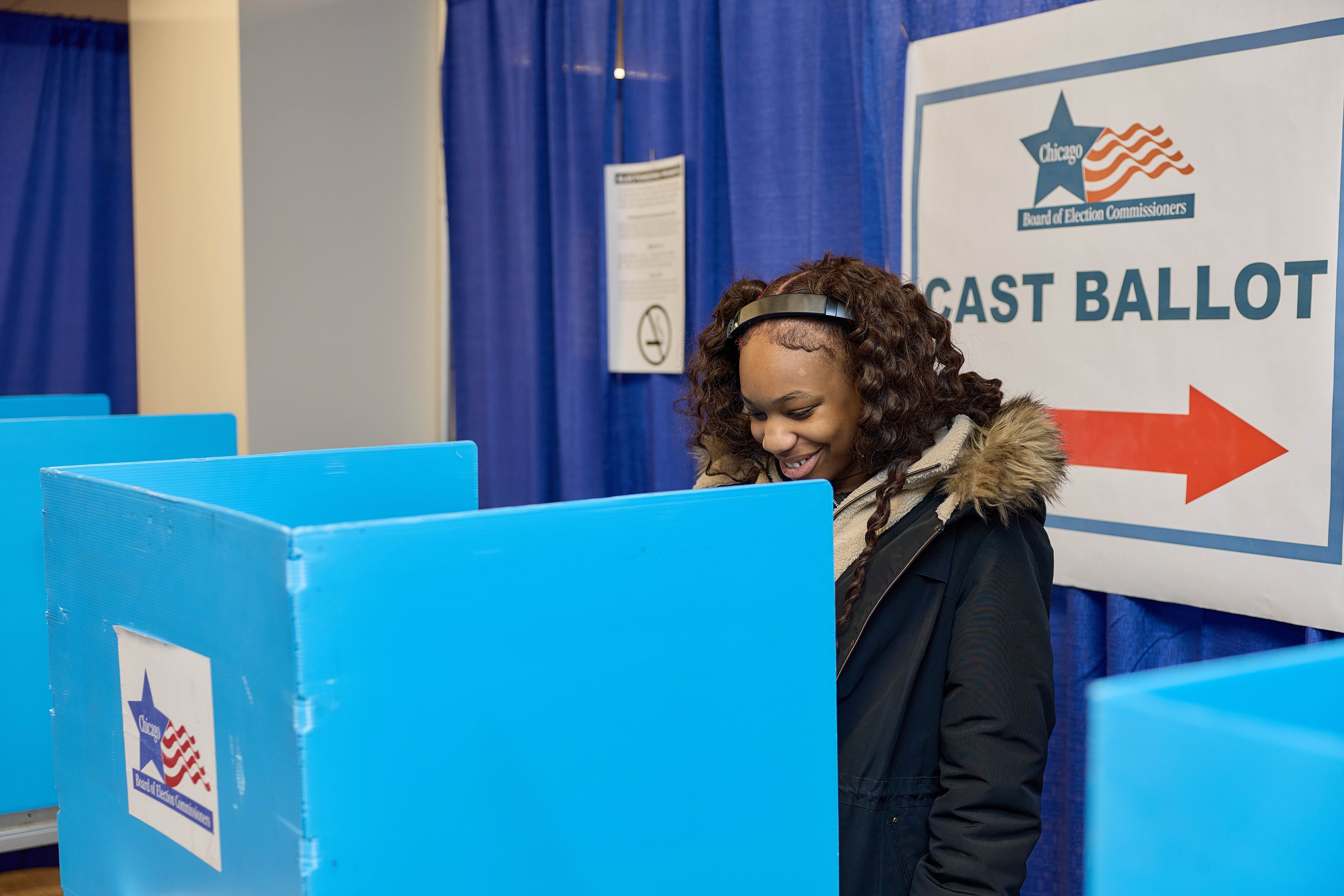 A Noble student stands behind a blue voting booth, smiling.