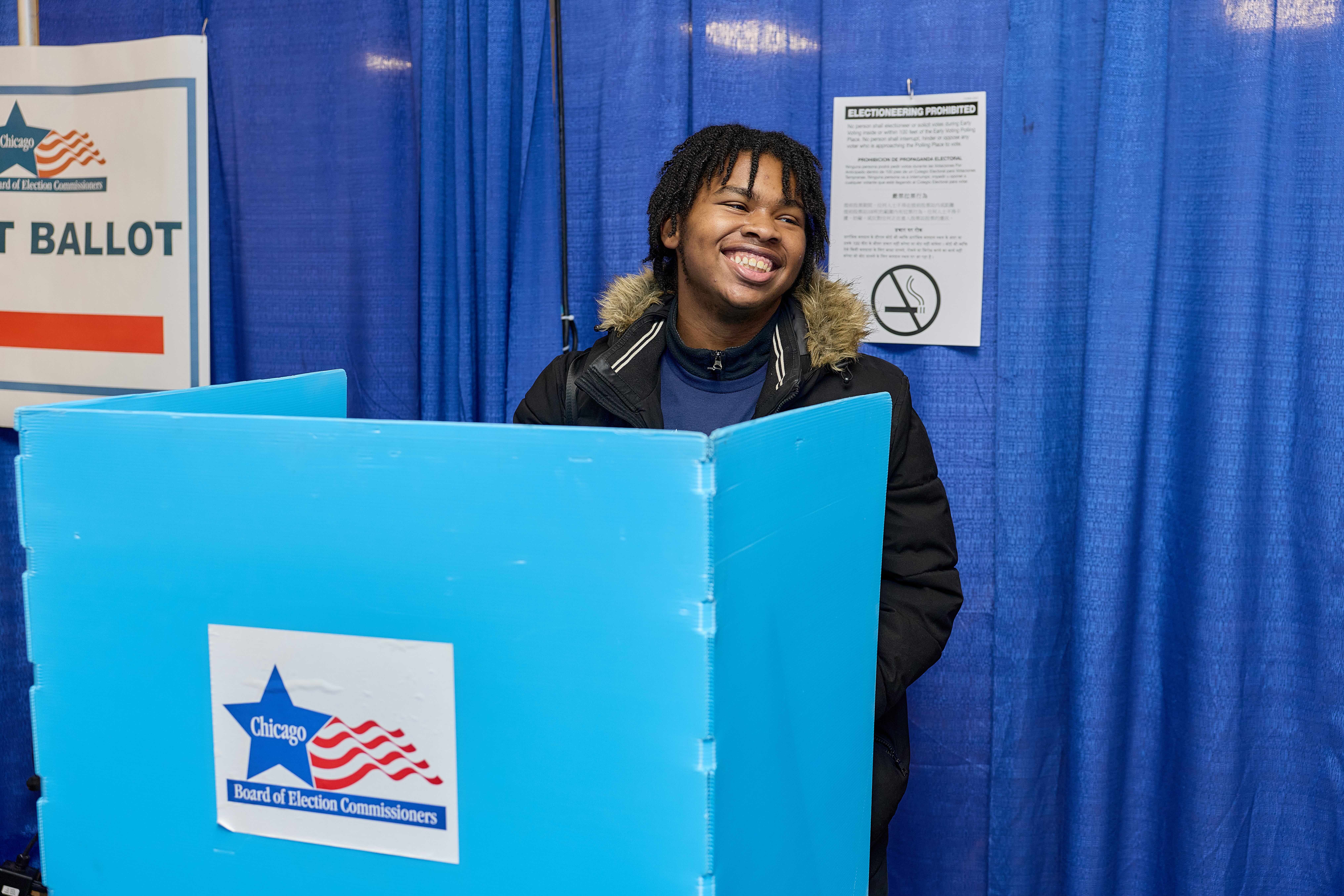 A Noble sutdent stands behind a blue voting booth. He is smiling wide, looking over to the side.