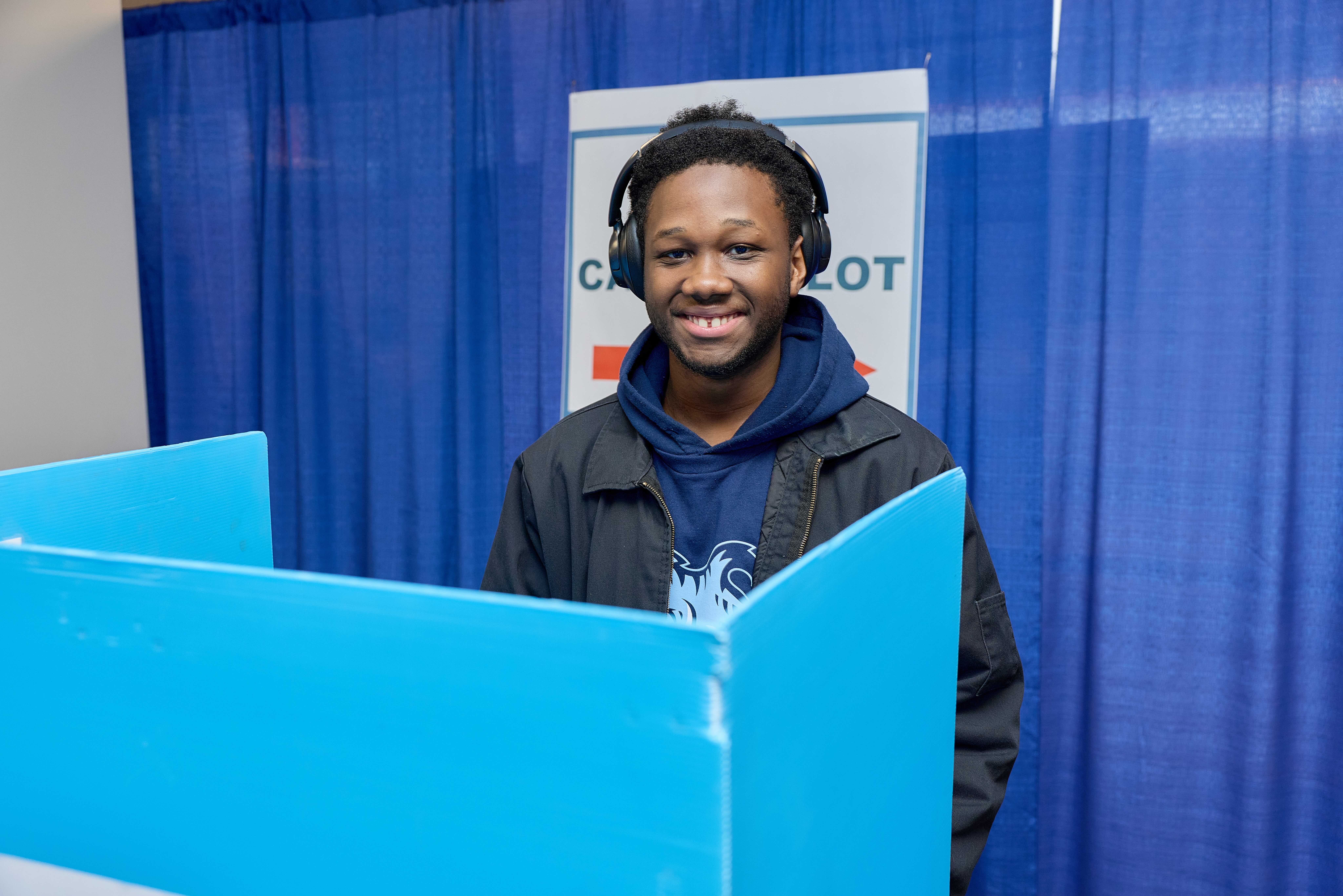 A Baker College Prep student stands behind a voting booth, smiling brightly.