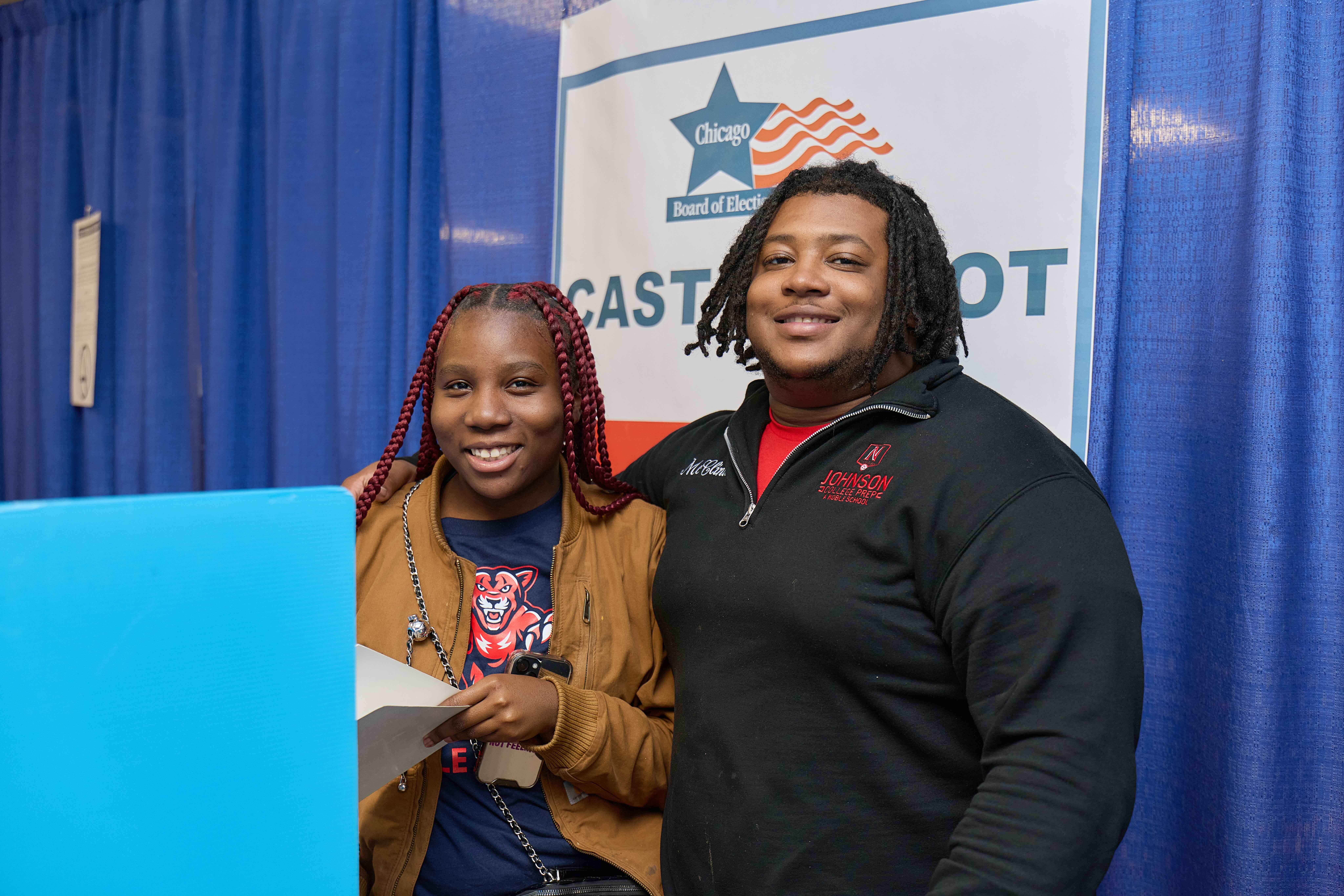 Two Johnson College Prep students pose for a photo behind the blue voting booth. They're smiling and the girl in front of the booth holds her voting papers in hand.